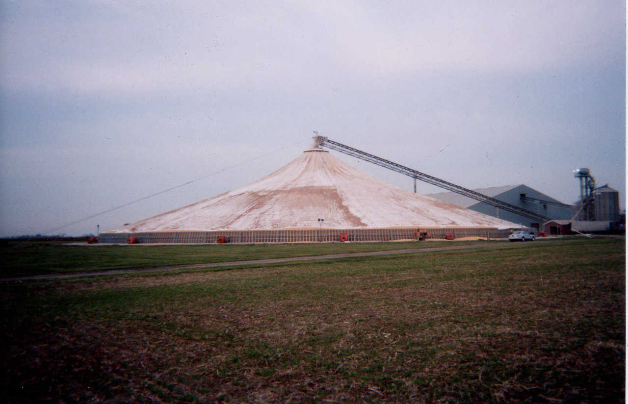 Grain is conveyed to the top of the tower via a conveyor, then dropped down the tower. As grain begins piling under the fabric cover, the lifting ring lifts it. When the fabric reaches its capacity, a cover is fastened over its central opening. Result: A cone-shaped, fabric structure full of protected, stored grain.
